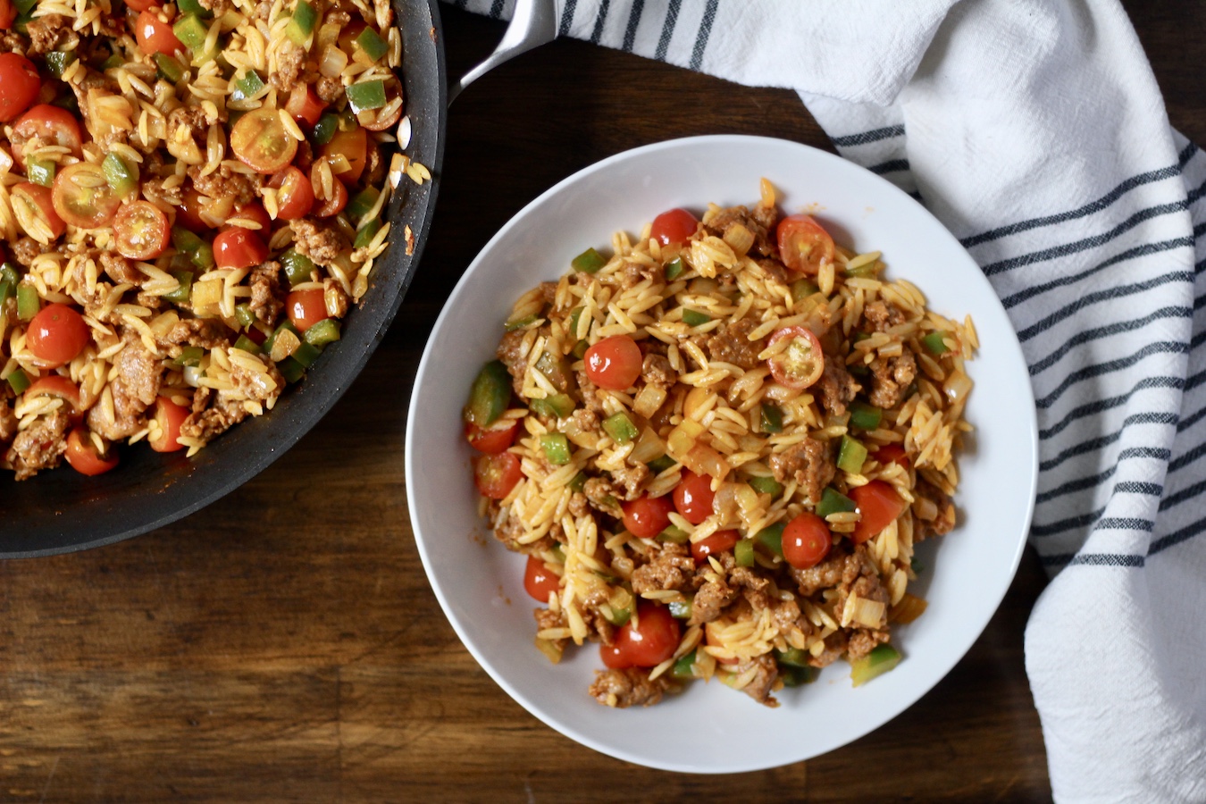 A white bowl with sausage and veggie orzo on a wooden table next to a skillet with sausage and veggie orzo and a white and blue towel at the top.