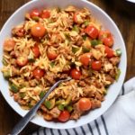 One large white bowl of sausage and veggie orzo with a fork in the bowl on a wooden table with a white and blue striped dish towel to the left of the photo.
