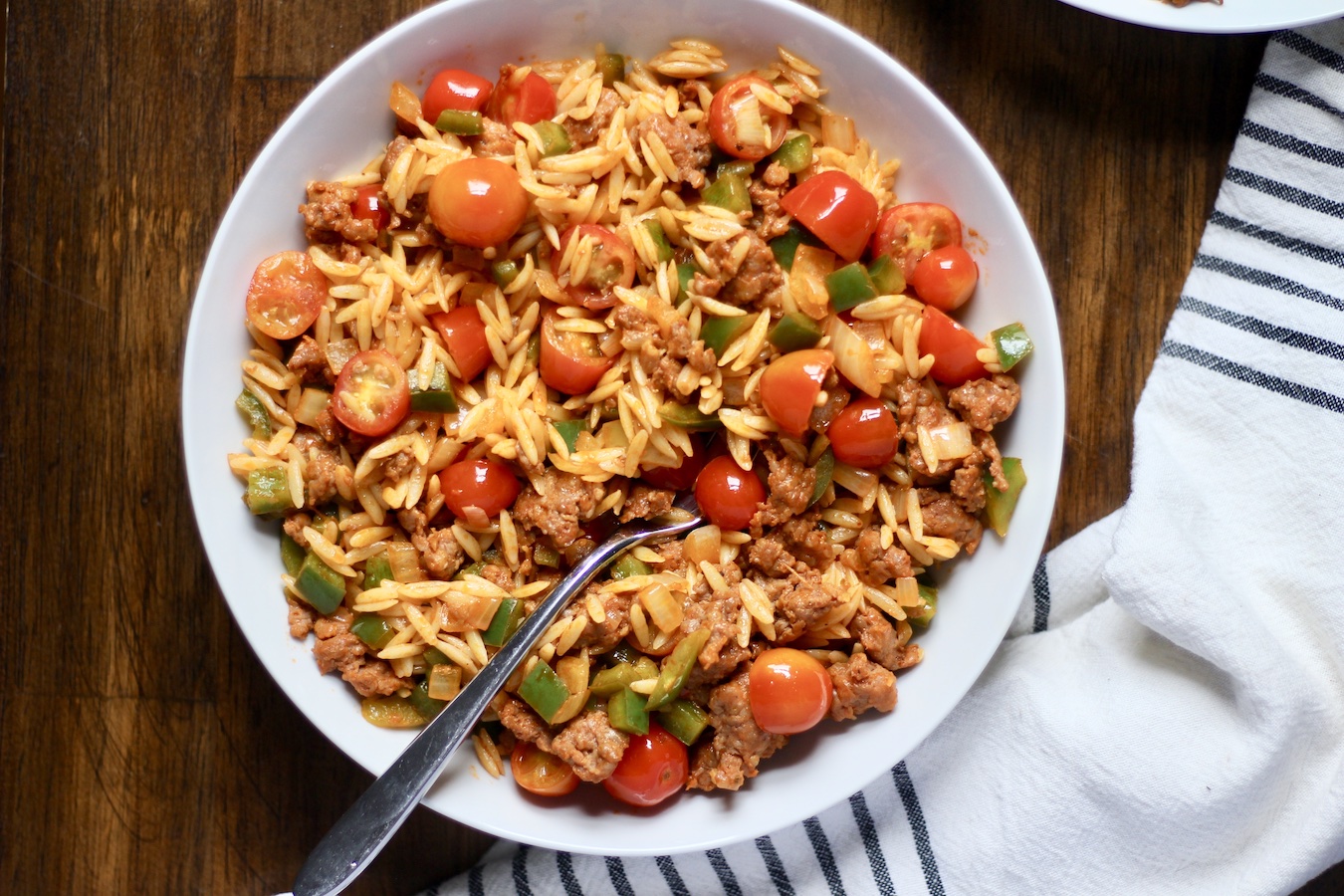 One large white bowl of sausage and veggie orzo with a fork in the bowl on a wooden table with a white and blue striped dish towel to the left of the photo.