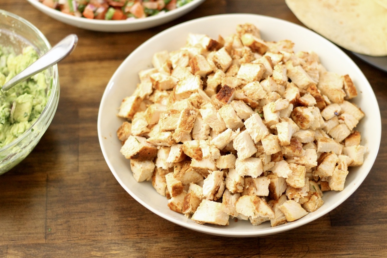 A white bowl of southwest chicken next to a bowl of guacamole on a wooden table.