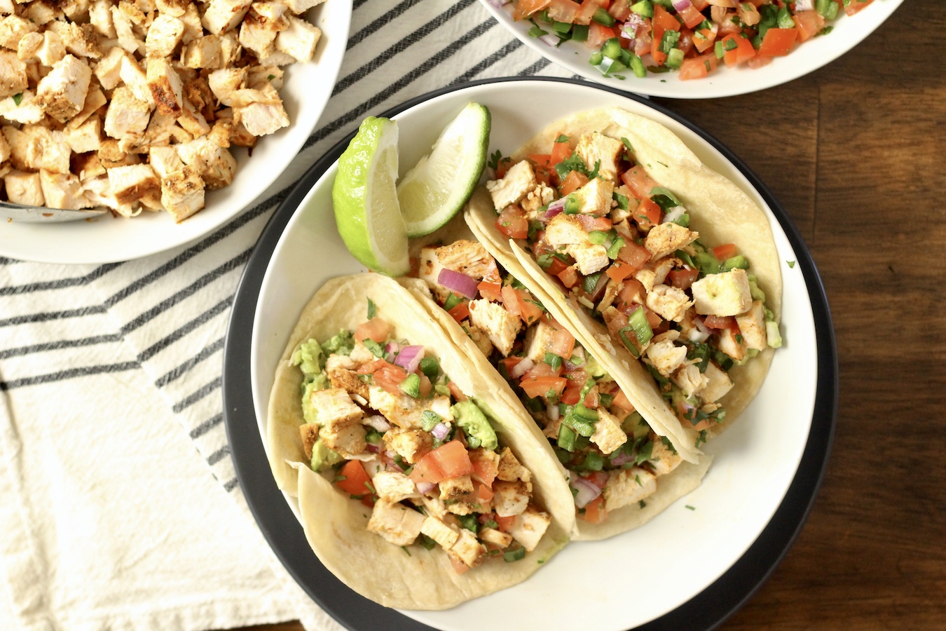 Three chicken tacos in a white bowl on a blue plate with chicken and pico de gallo in the back.