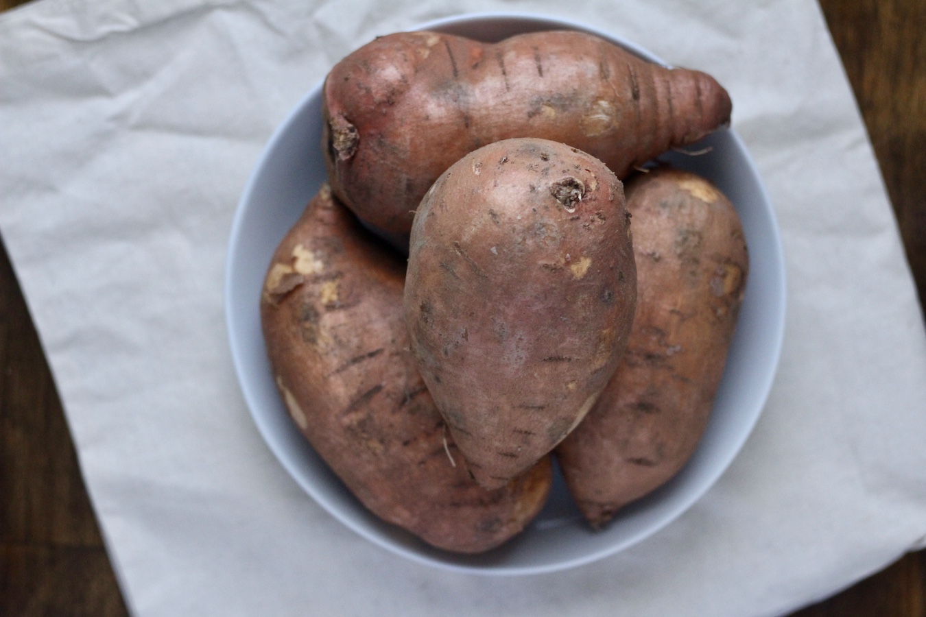 White bowl of uncooked sweet potatoes on a canvas bag on a wooden table.