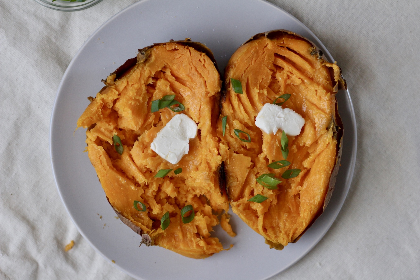 Sweet potato cut lengthwise with unsalted vegan butter and green onion on a white plate on a canvas bag.
