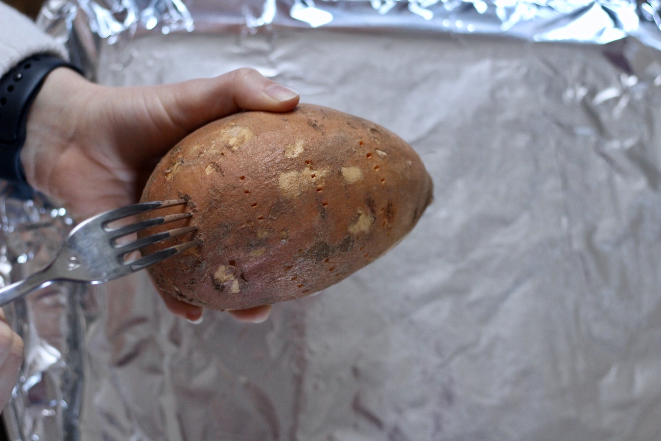 Two hands poking holes in a sweet potato over a foil lined baking sheet.