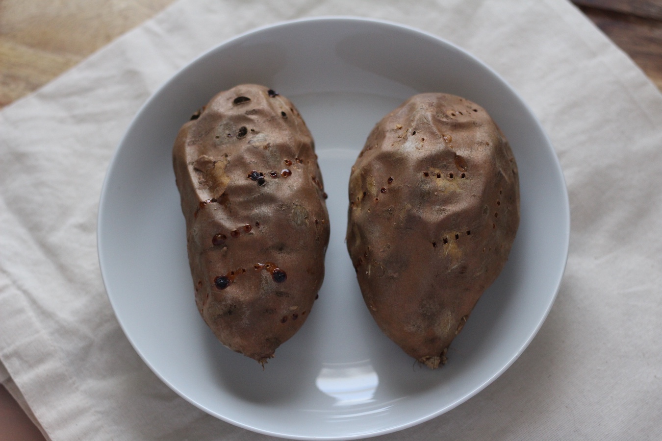 Two oven baked sweet potatoes in a white bowl on a canvas bag on a wooden counter.