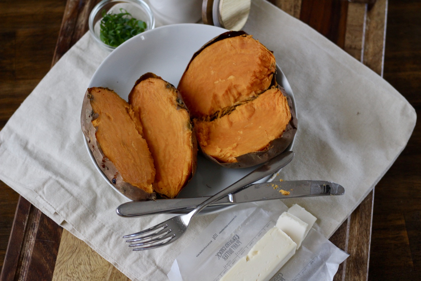 Lengthwise cut sweet potatoes in a white bowl on a canvas bag with a knife and fork in the front with unsalted vegan butter.
