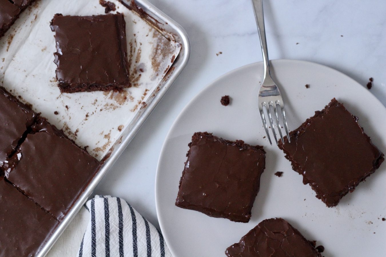 Three squares of cake on a white plate with fork and a sheet pan with more cake cut into squares.