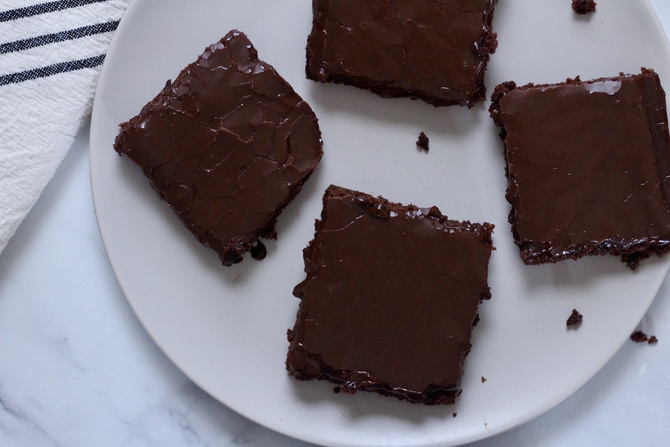 Four cut squares of Texas Sheet Cake on a white plate on a marble counter with a white and blue striped dish towel to the left.