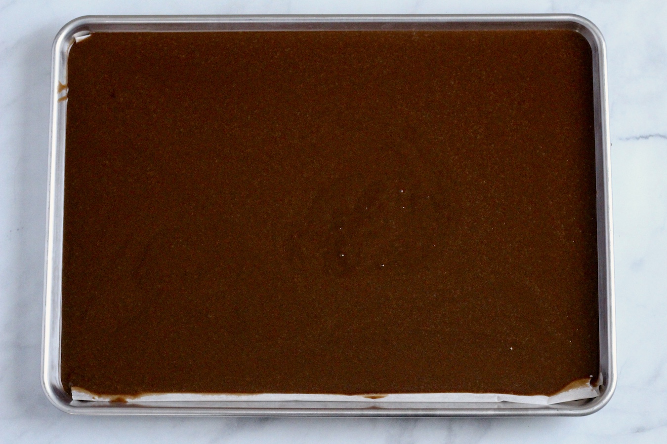 Chocolate sheet cake batter in a sheet pan on a marble counter.