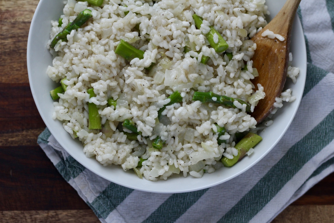 Asparagus risotto in a white bowl on a green and white striped towel on a wooden counter.