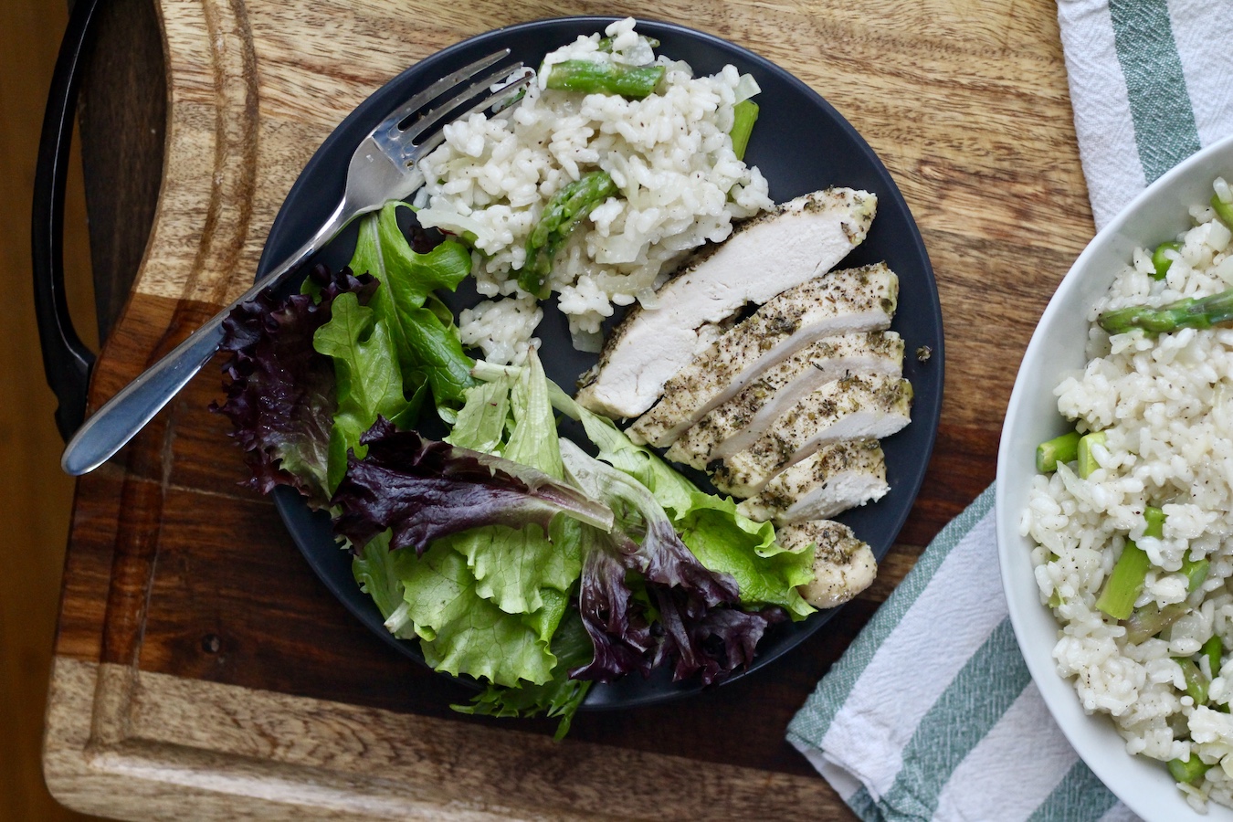 Chicken, salad, and asparagus risotto on a plate with a fork on a wooden counter.