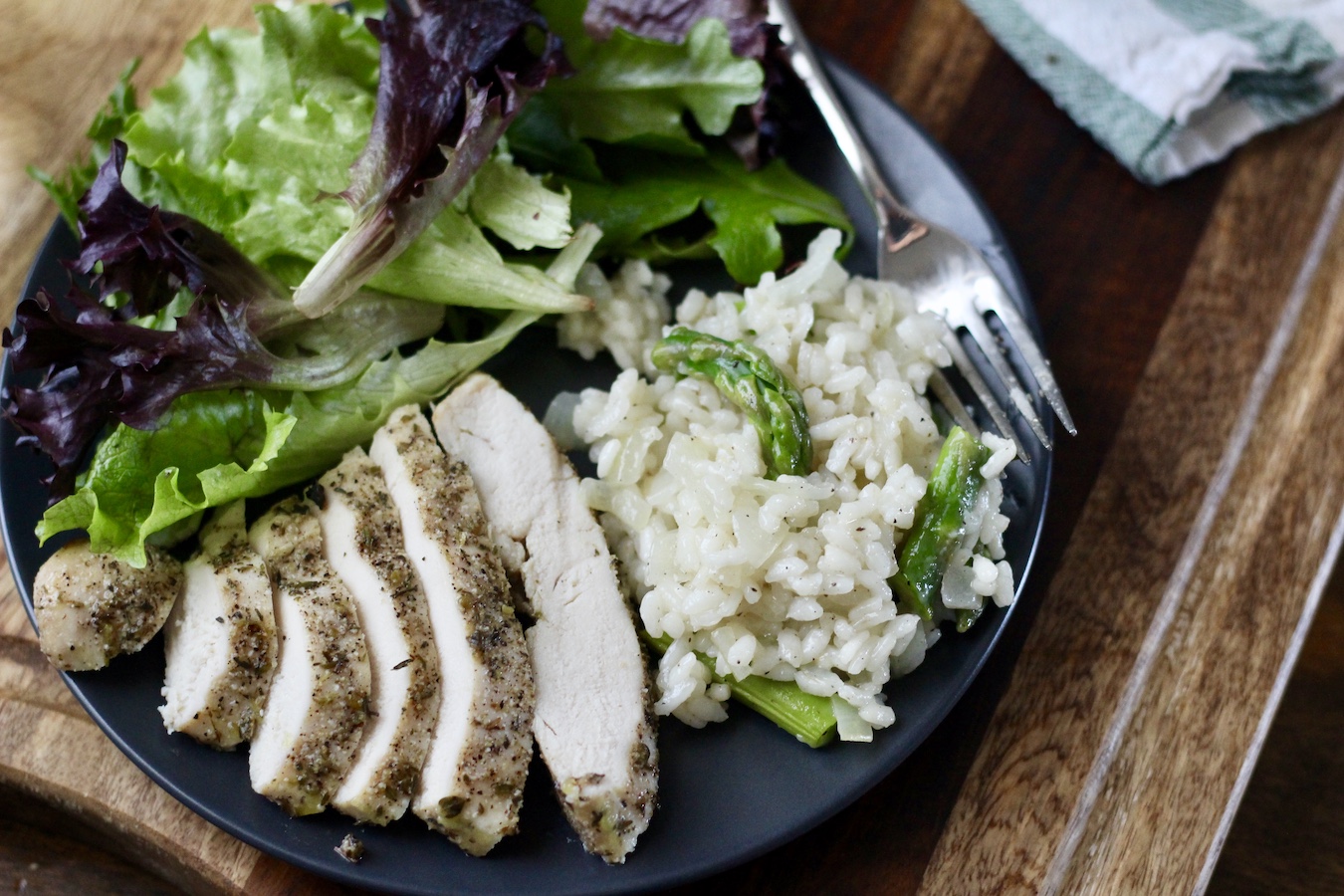 Chicken, salad, and asparagus risotto on a plate with a fork on a wooden counter.