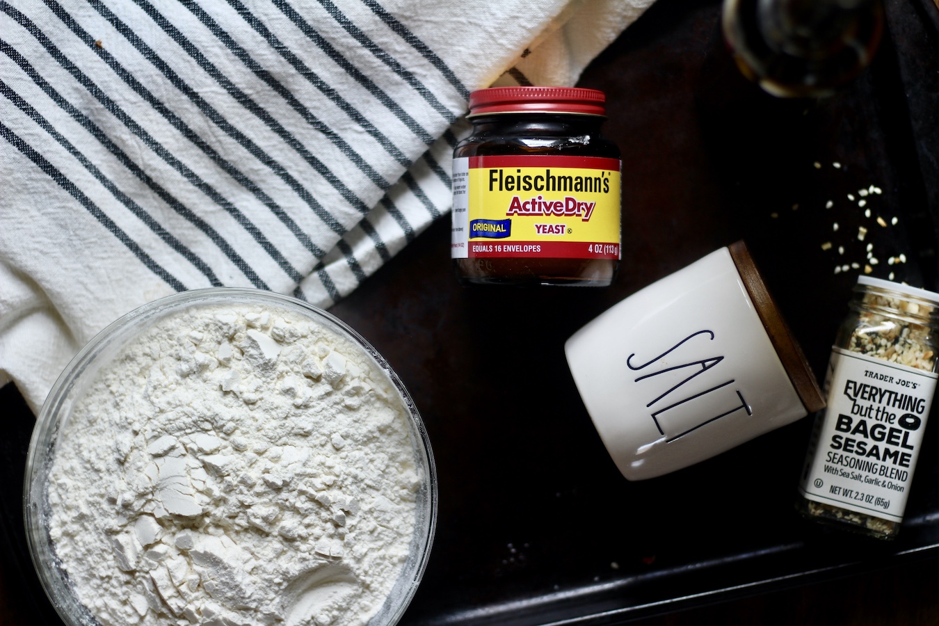 Flour in a glass bowl on the left, yeast in a container in the middle, a salt jar to the right, and a jar of everything but the bagel seasoning blend to the far right on a cookie sheet with a white and blue dish towel.