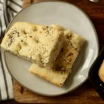 Two slices of breakfast focaccia on a small cream plate with a white and blue striped dish towel to the left.