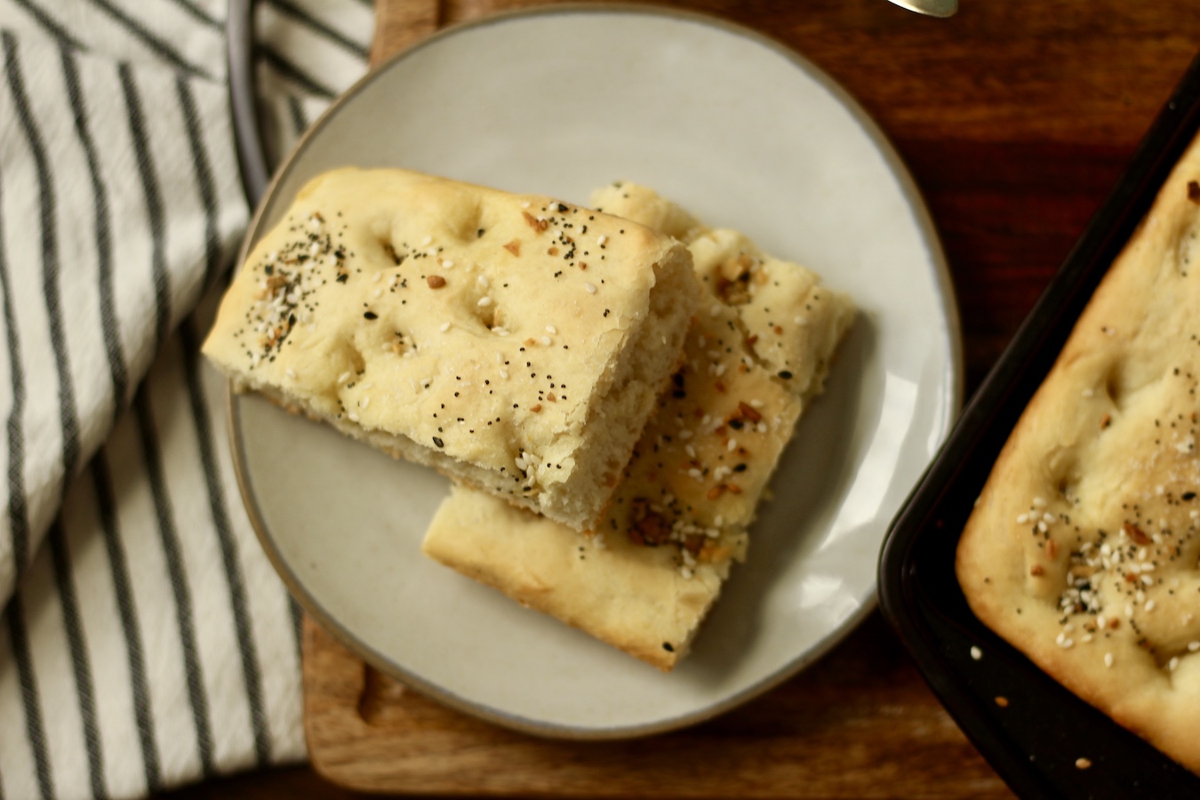 Two slices of breakfast focaccia on a small cream plate with a white and blue striped dish towel to the left.