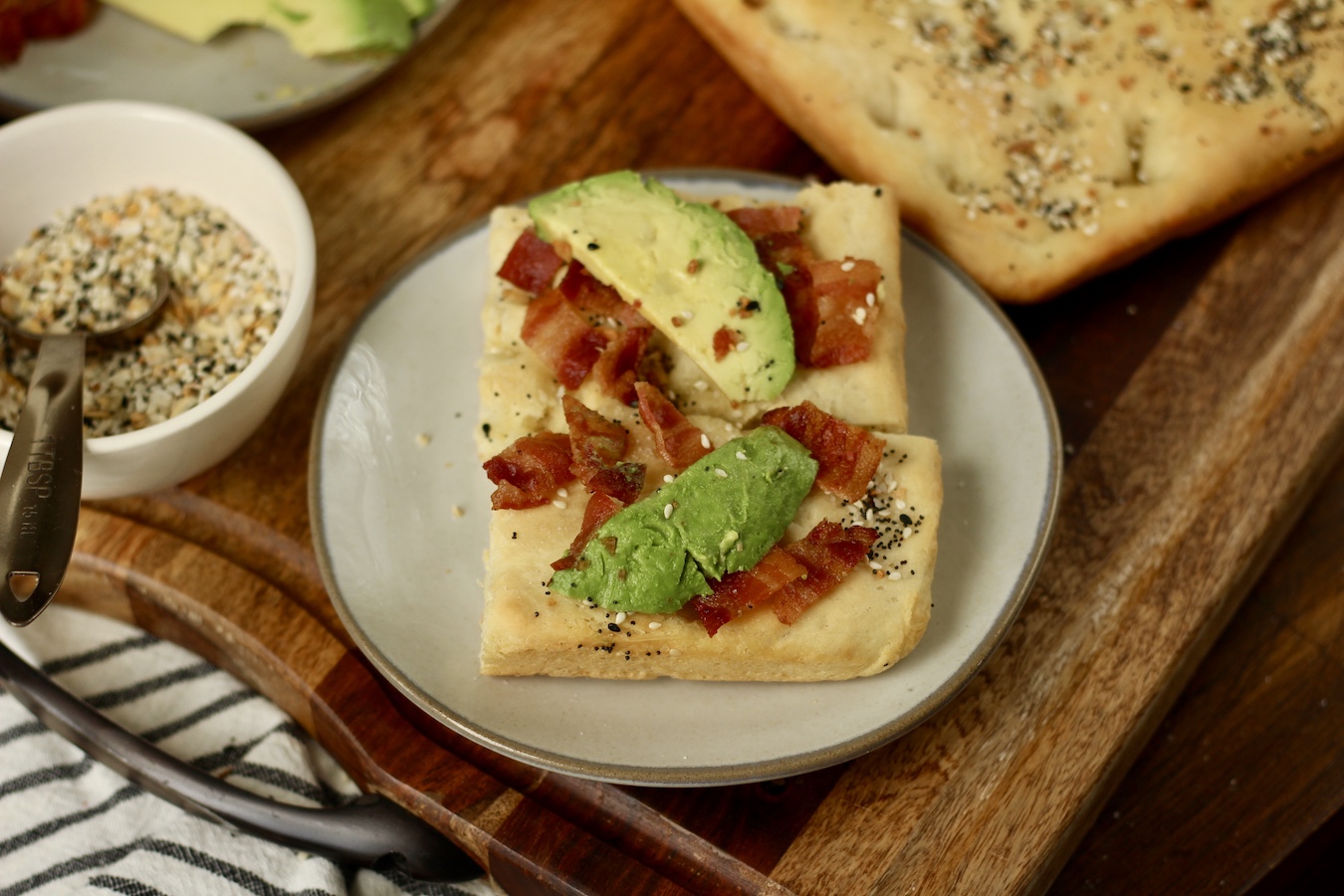 A cream plate with breakfast focaccia topped with bacon and avocado, a bowl of everything but the bagel seasoning to the left, and more focaccia to the right.
