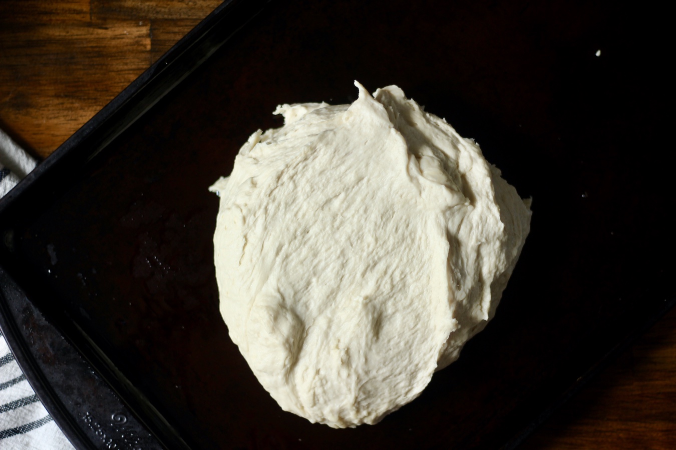Finished bread dough on a cookie sheet on a wooden table with a blue and white striped dish towel to the left.