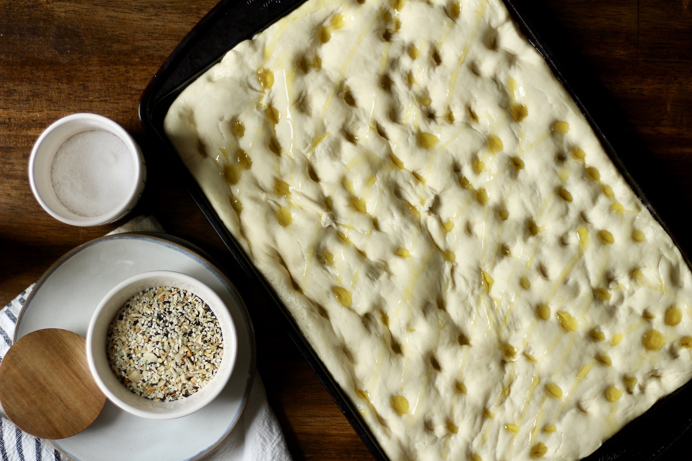 Breakfast focaccia on a baking sheet with holes pushed in and olive oil drizzled on top with salt and everything but the bagel seasoning to the left.