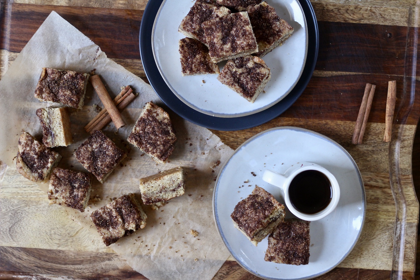 Cinnamon crumble bars on the left side of the cutting board with more stacked on a plate in the back and two stacked on a plate in front of a cup of coffee.