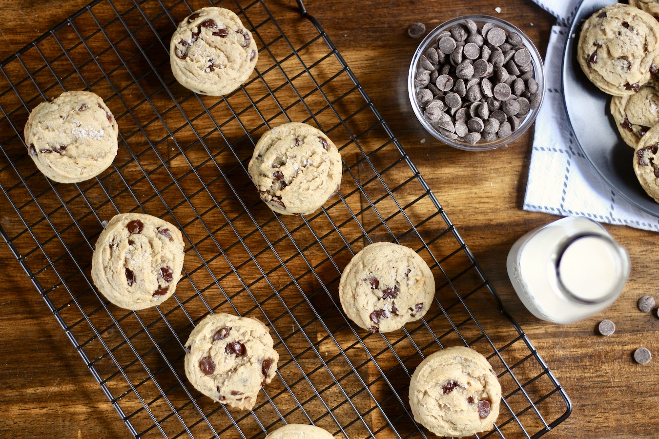 Egg free chocolate chip cookies on a cooling rack on a wooden table with chocolate chips in a small bowl and a small jug of nut milk on the right.