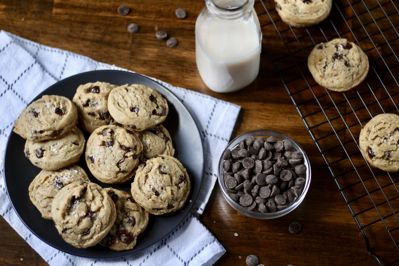 A blue plate of egg free chocolate chip cookies on a white dish towel on a wooden table with a small bowl of chocolate chips and a jug of nut milk in the middle and a cooling rack of cookies on the right.
