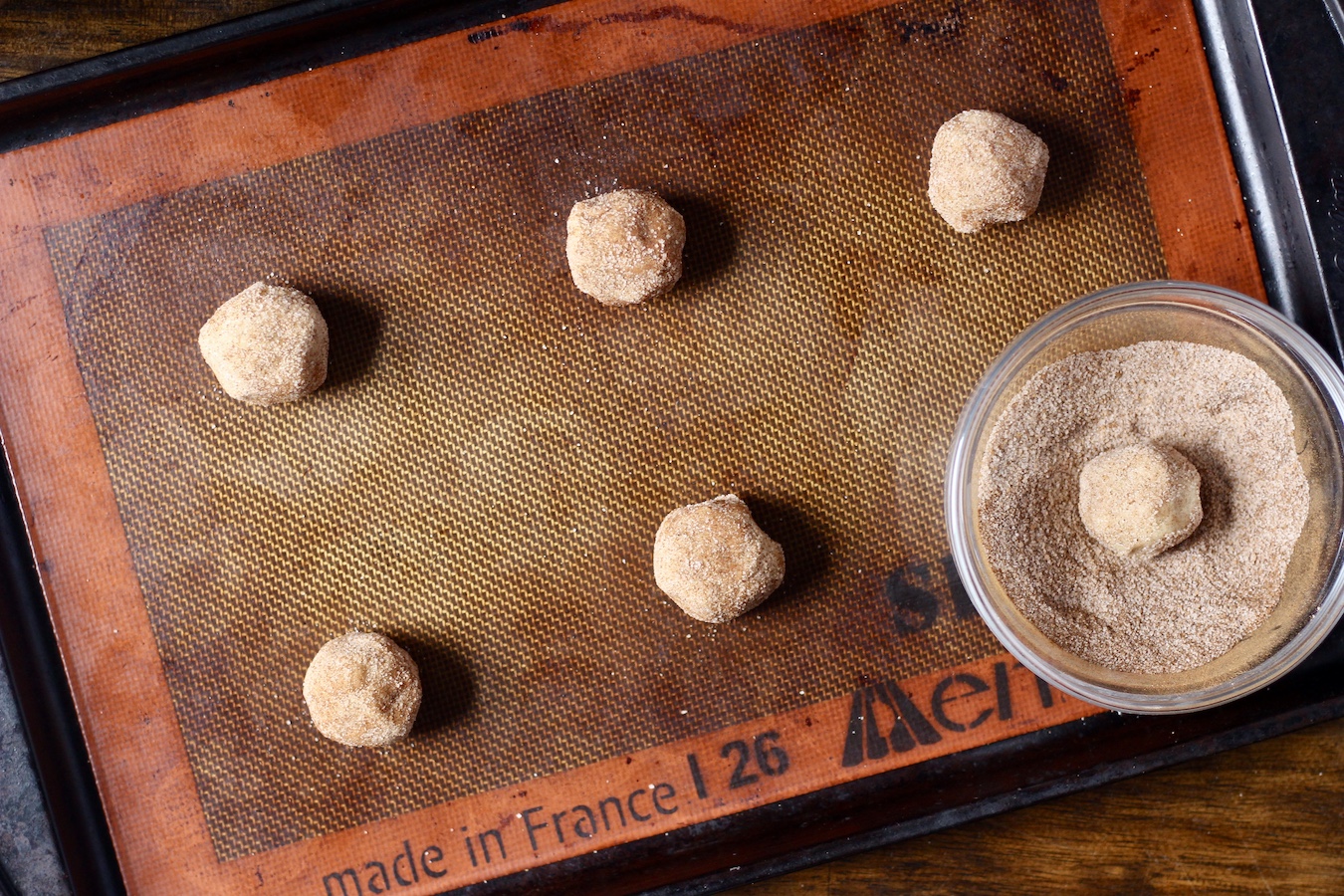 Six dough balls on a cookie sheet with a bowl of cinnamon sugar in the right corner.