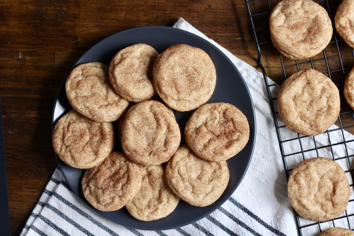 snickerdoodle cookies on a blue plate and cooling rack on a white and blue towel on a wooden table.