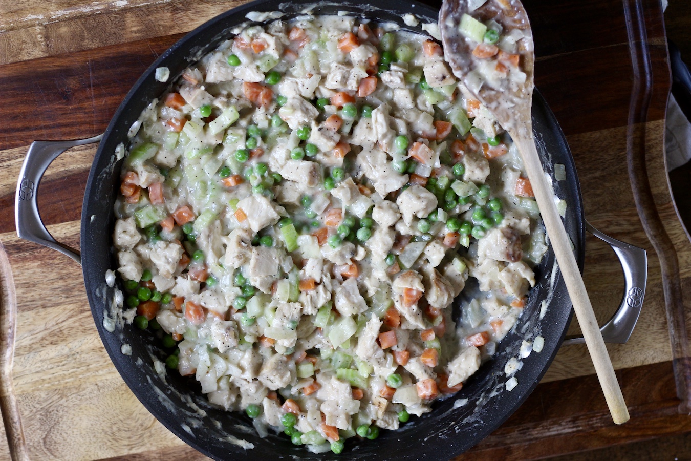 The chicken pot pie filling in a skillet on a wooden cutting board with a wooden spoon.