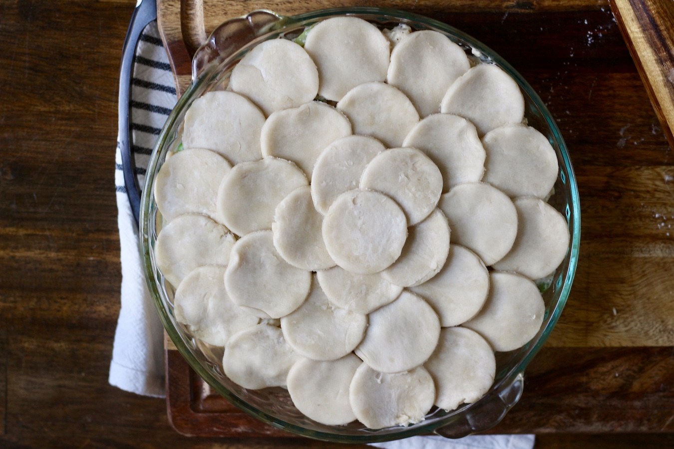 The cut pastry dough in circles on top of the chicken pot pie filling in the pie pan on a wooden cutting board.