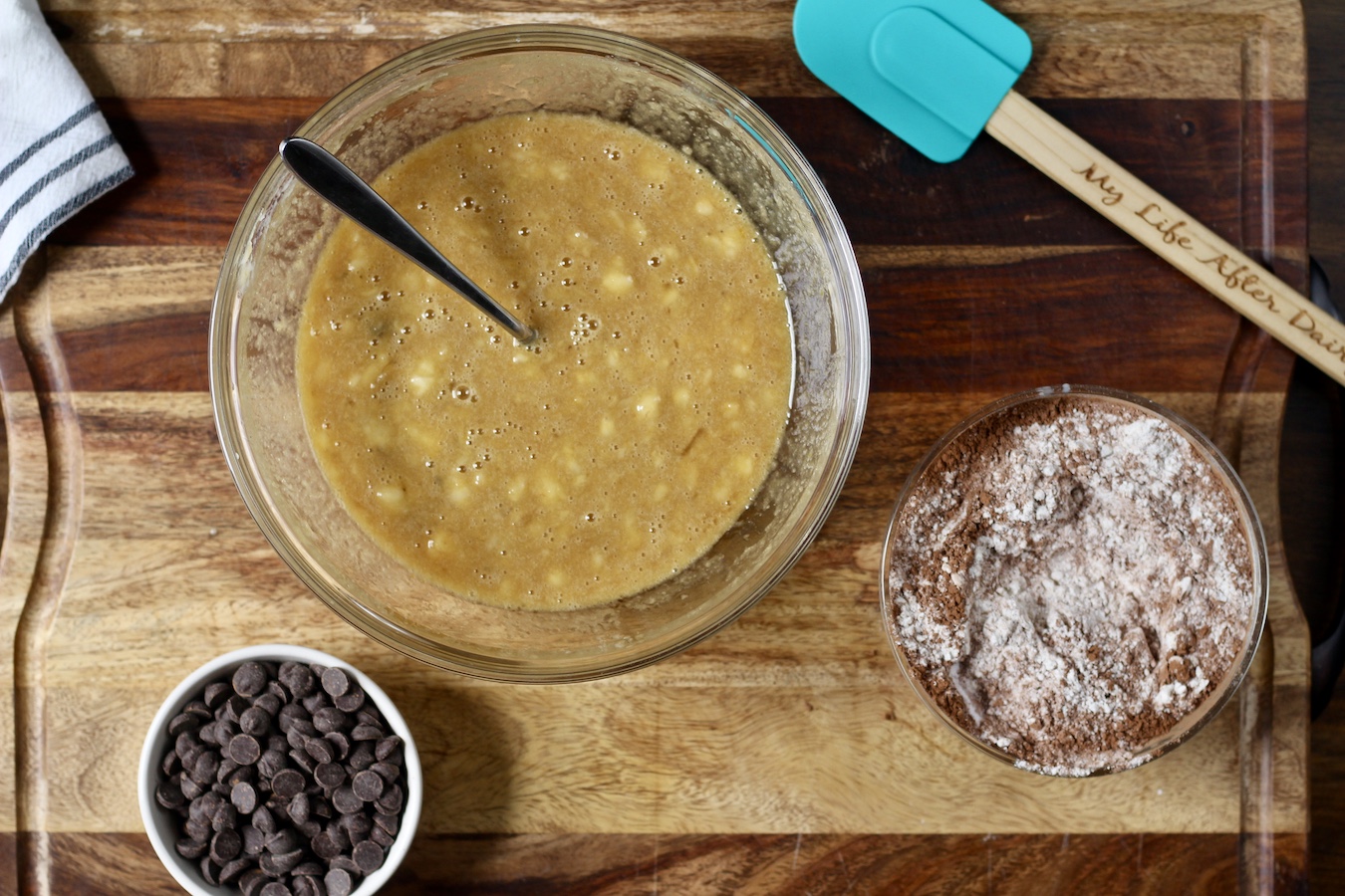 A bowl with chocolate chips, a large bowl with the wet ingredients, and a small bowl with the dry ingredients on a wooden cutting board with a rubber spatula.