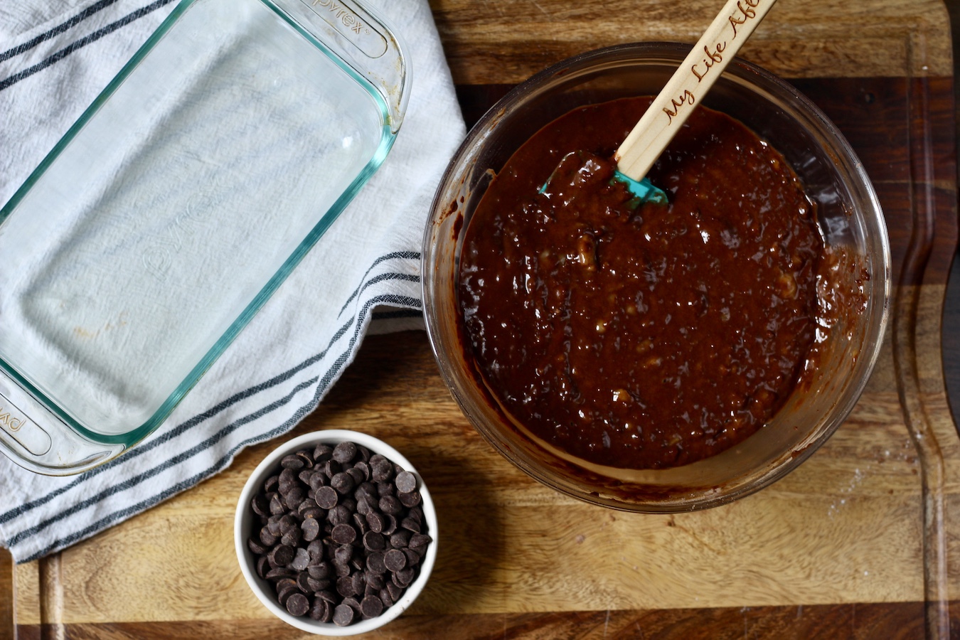 Chocolate banana bread batter in a large bowl with a rubber spatula on a wooden counter with a small bowl of chocolate chips and a bread pan to the left.