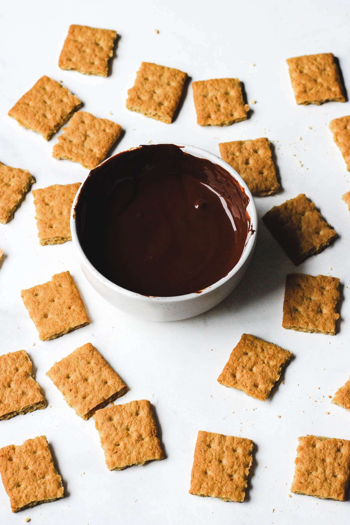 A small white bowl of melted dark chocolate with graham cracker squares all around the bowl.