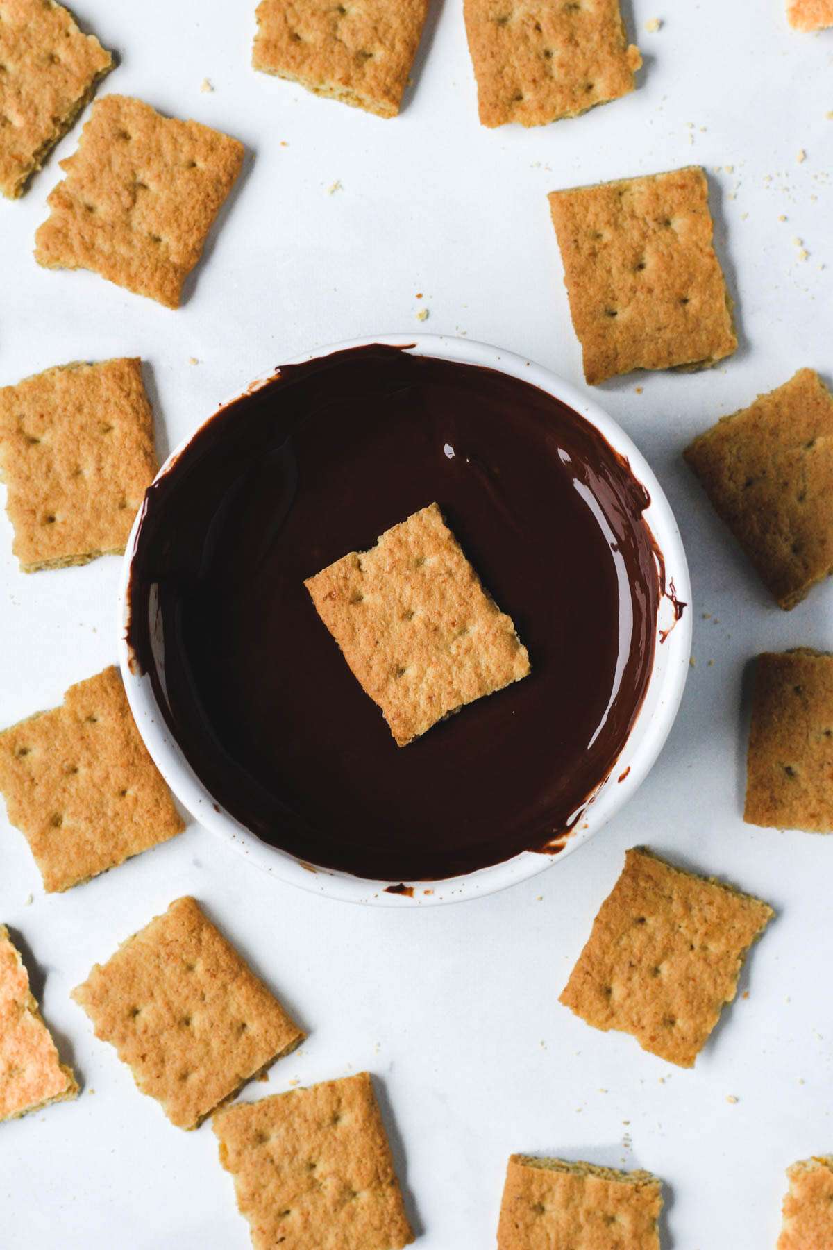 A top down photo of a small white bowl with dark chocolate and a graham cracker being dipped in the bowl.