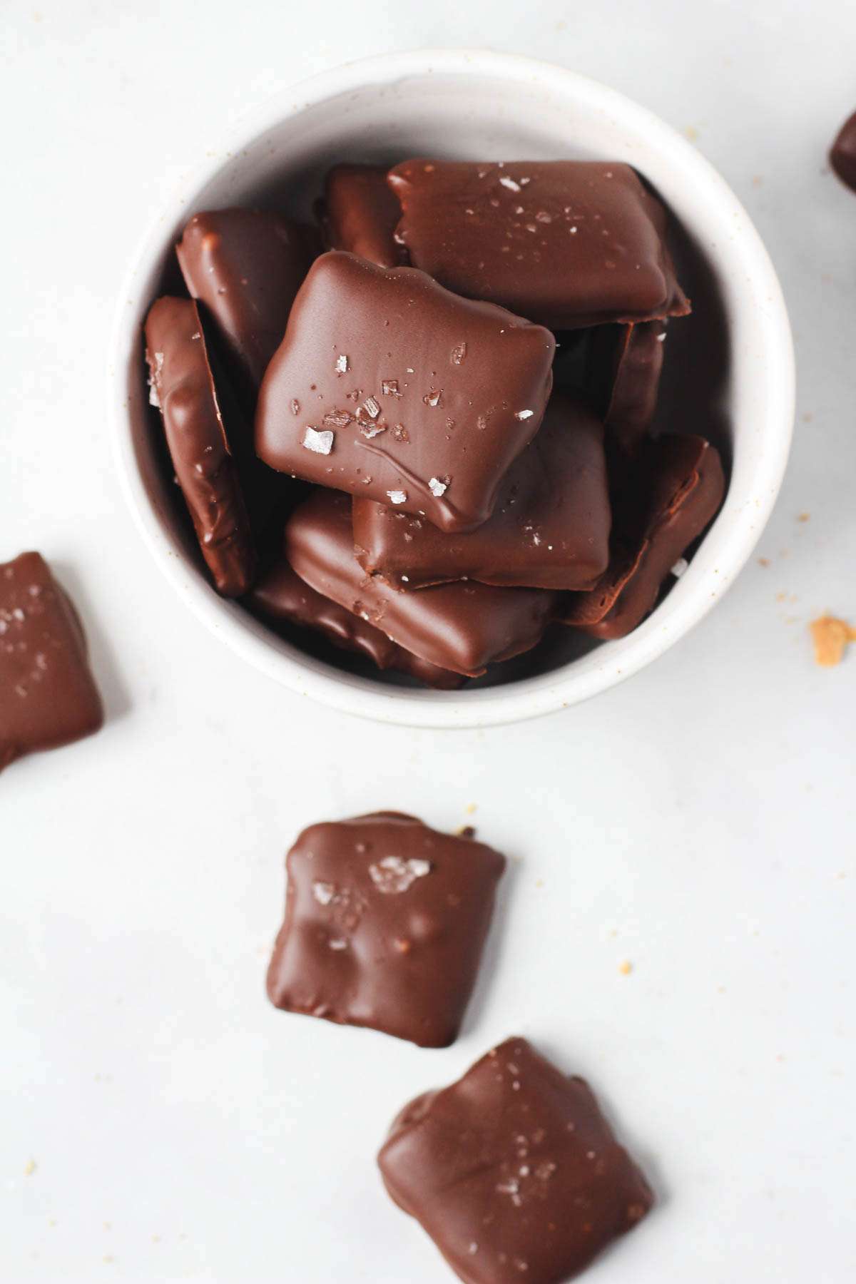 A small white bowl filled with dairy-free dark chocolate dipped graham crackers sprinkled with flaky sea salt and a few graham crackers sprinkled on the counter.