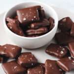 A white bowl filled with dark chocolate dipped graham crackers with sea salt on a white plate with more chocolate coated graham crackers.