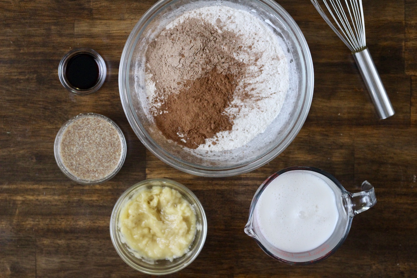 Small bowls with ingredients on a wooden table with a whisk to the right.