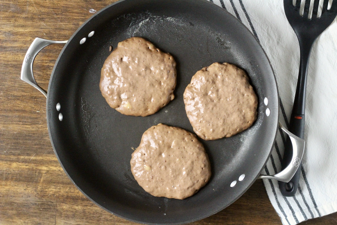 A nonstick pan with three pancakes on a wooden table with a dish towel and a spatula.