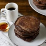 A stack of Chocolate Protein Pancakes on a plate with a fork to the left, syrup, and a small cup of coffee on a dish towel.