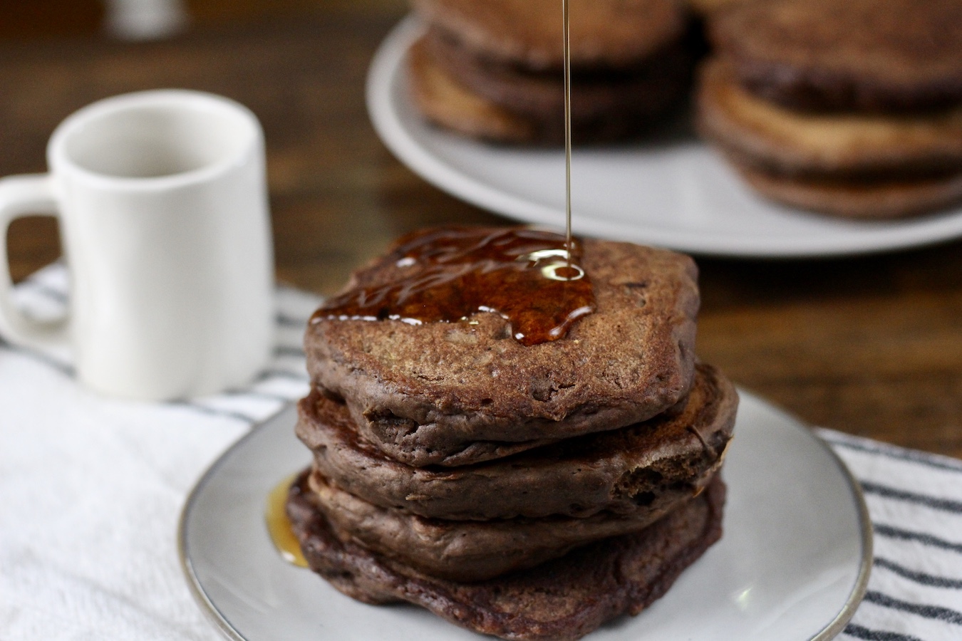 A stack of Chocolate Protein Pancakes on a plate with syrup being poured on them and a small cup of coffee in the back.