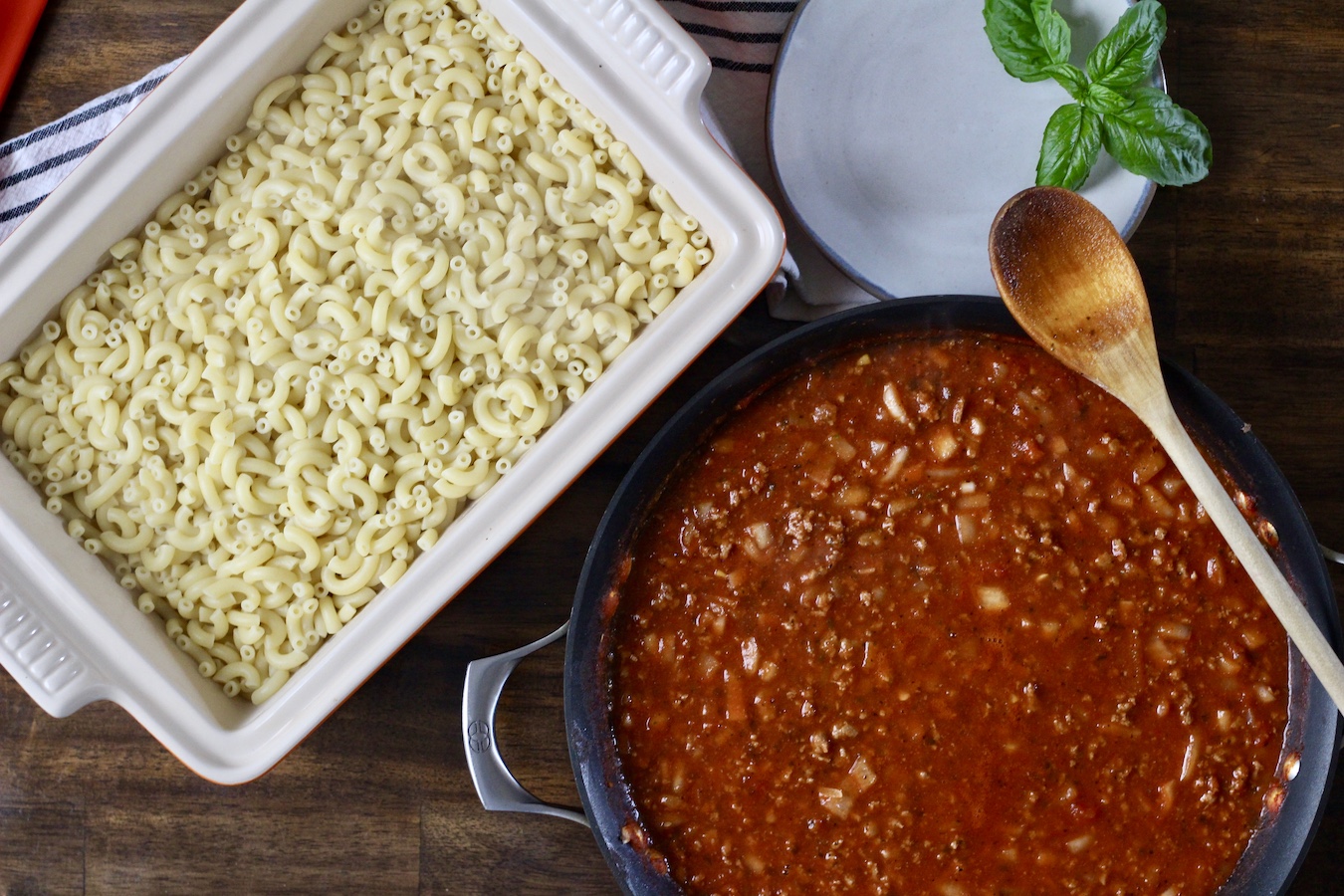 A baking dish with cooked pasta and a skillet of meat sauce with a wooden spoon.
