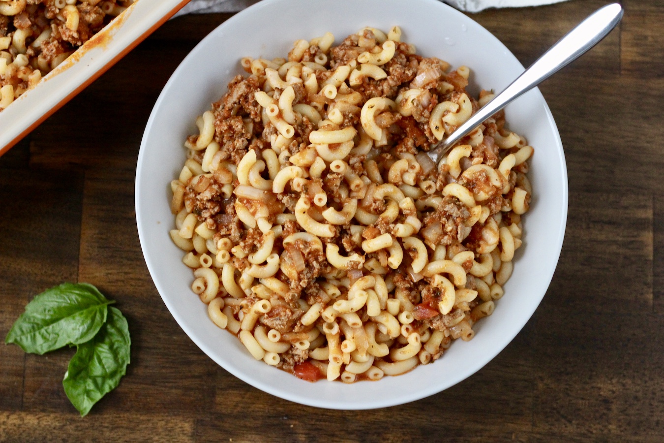 A white bowl with homemade hamburger helper, a spoon, and some basil to the bottom left.
