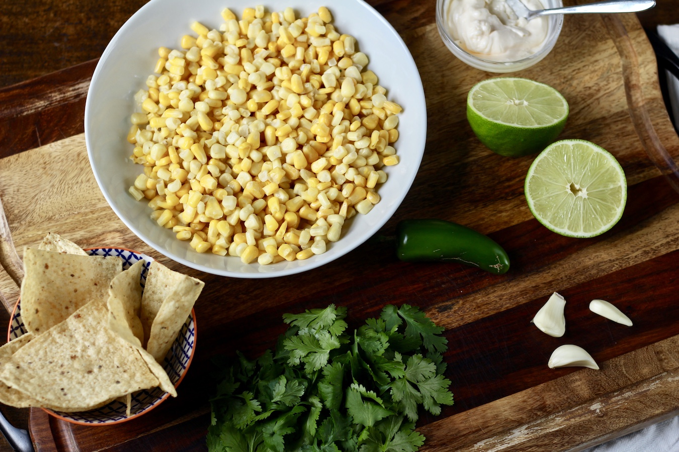 Corn in a white bowl with a small bowl of mayo, limes, garlic pods, a jalapeño, cilantro, and a bowl of tortilla chips on a wooden cutting board.