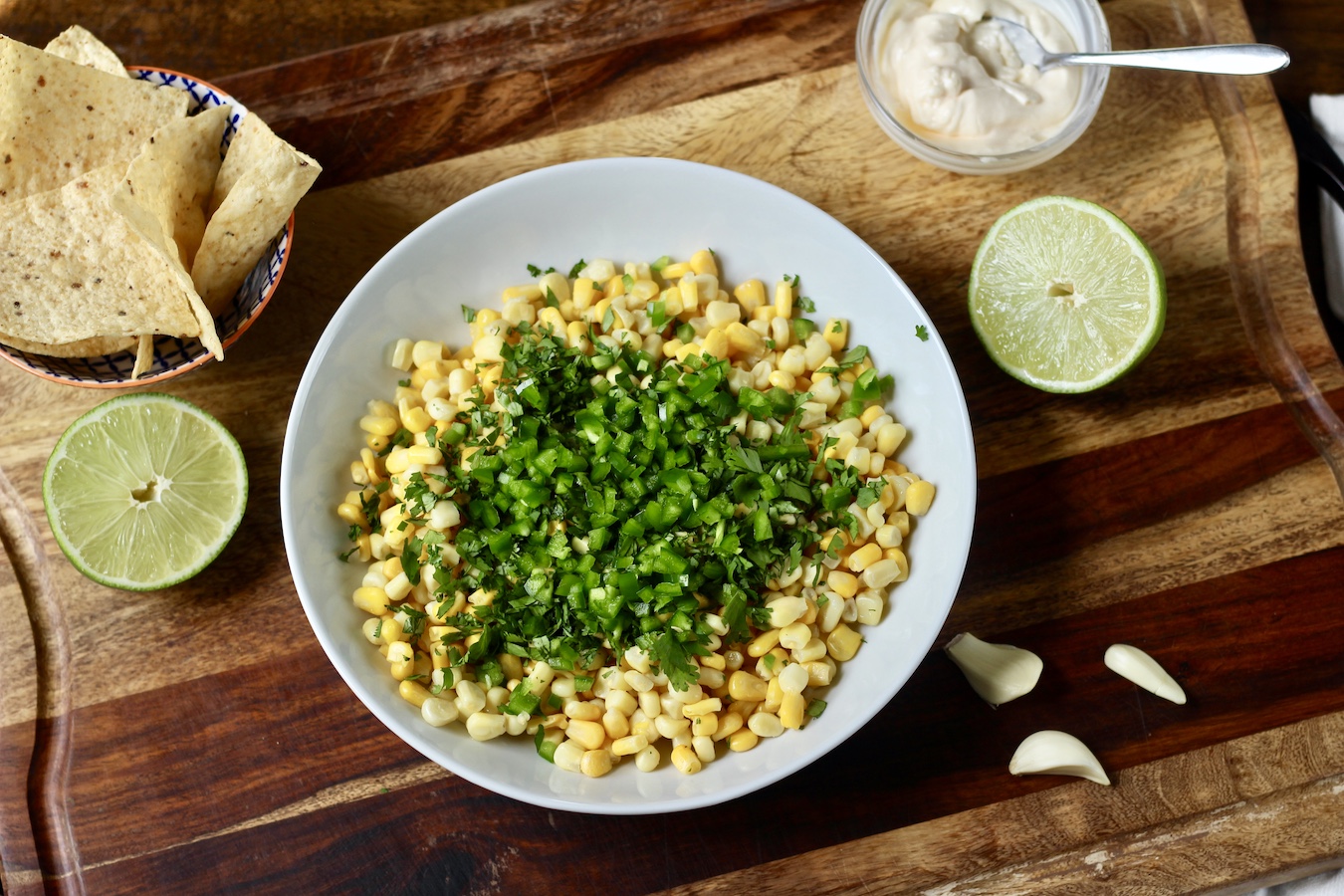 Corn in a white board with chopped jalapeño and cilantro on a wooden cutting board with a bowl of tortilla chips, limes, a small bowl of mayo and garlic pods.