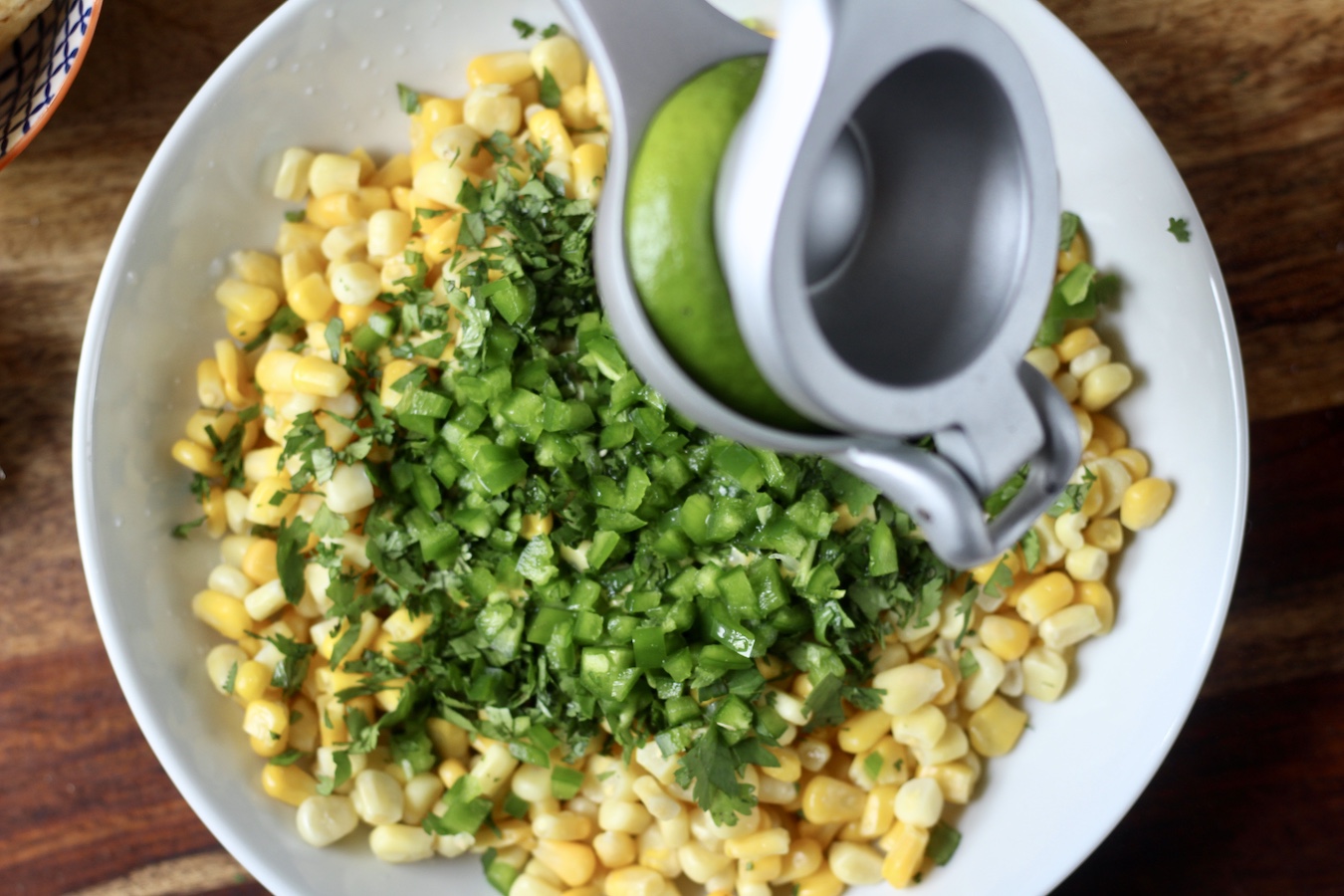 A lime being juiced into a bowl with corn, jalapeño, and cilantro on a wooden cutting board.