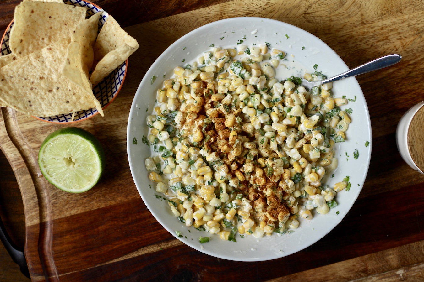 A bowl of Mexican street corn dip with a spoon and a small bowl of tortilla chips and a lime on a wooden cutting board.