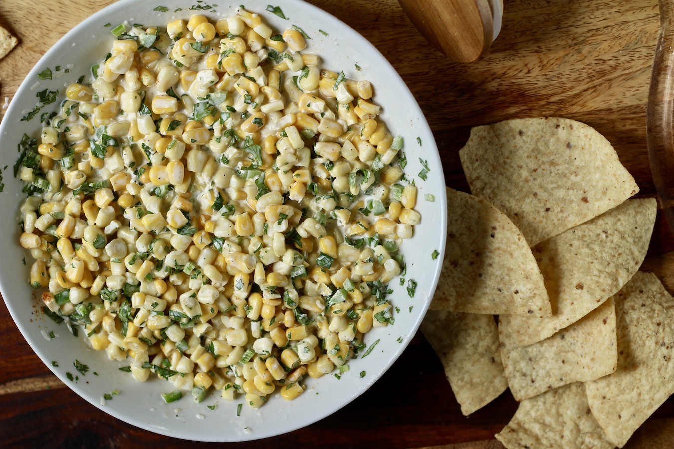 A white bowl with mexican street corn dip with a pile of tortilla chips on the right on a wooden cutting board.