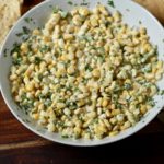 Large white bowl of mexican street corn dip on a wooden cutting board with tortilla chips on the side.