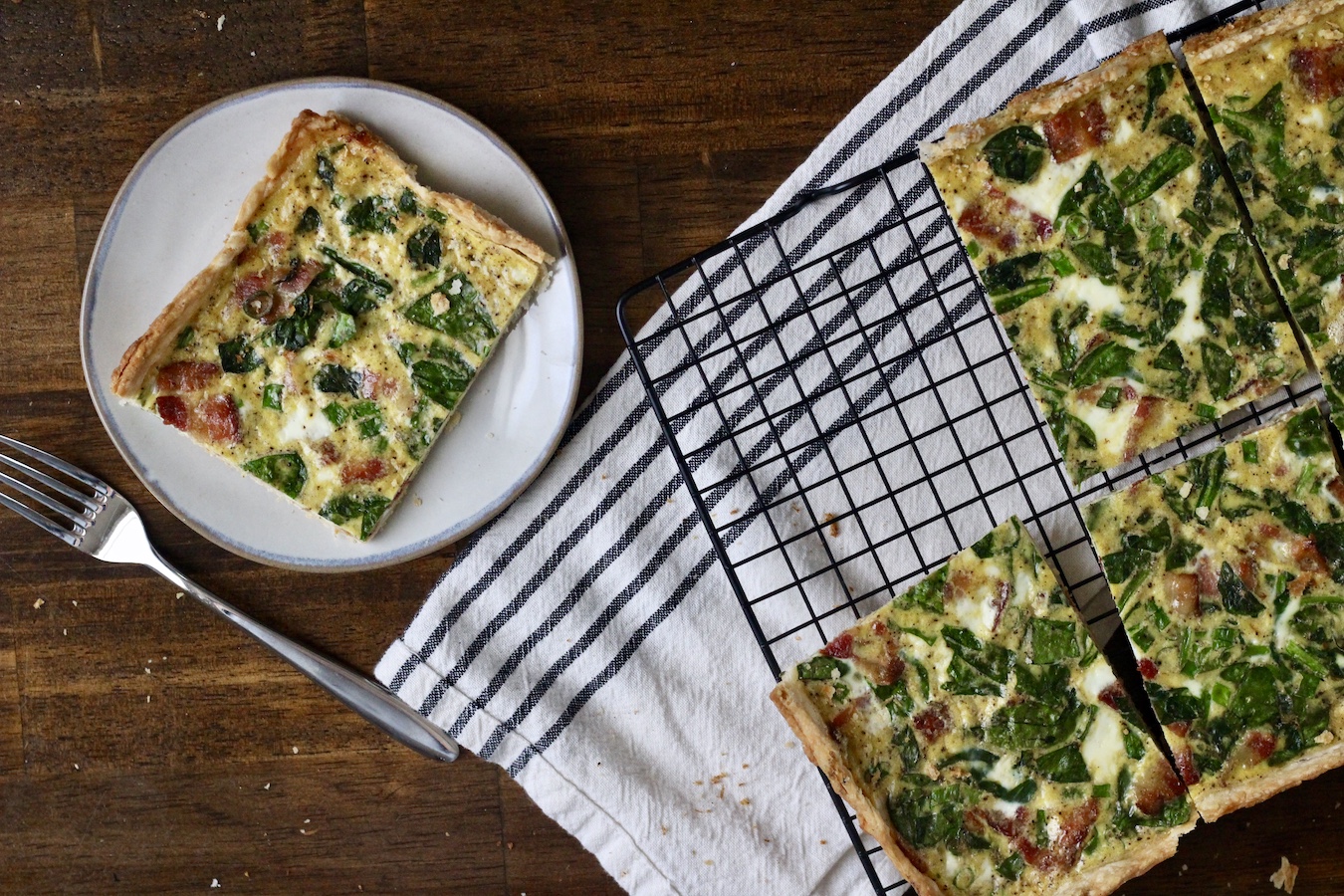 Slab quiche cut into squares on a cooling rack and a slice on a white plate with a fork to the left.