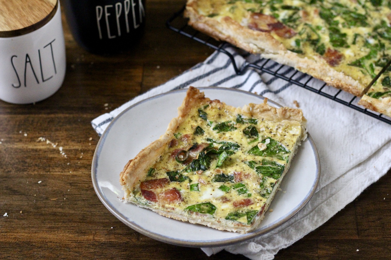 A square of slab quiche on a white plate with salt and pepper in the back.
