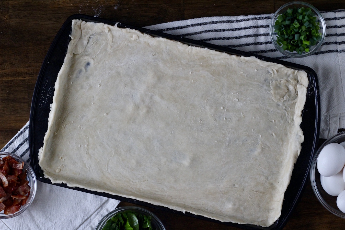 Pastry dough rolled out and on the baking sheet with eggs, bacon, and green onion around the side.